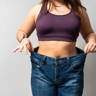 Woman wearing oversized jeans, celebrating successful weight loss journey through dedicated diet and workout, showcasing impressive transformation